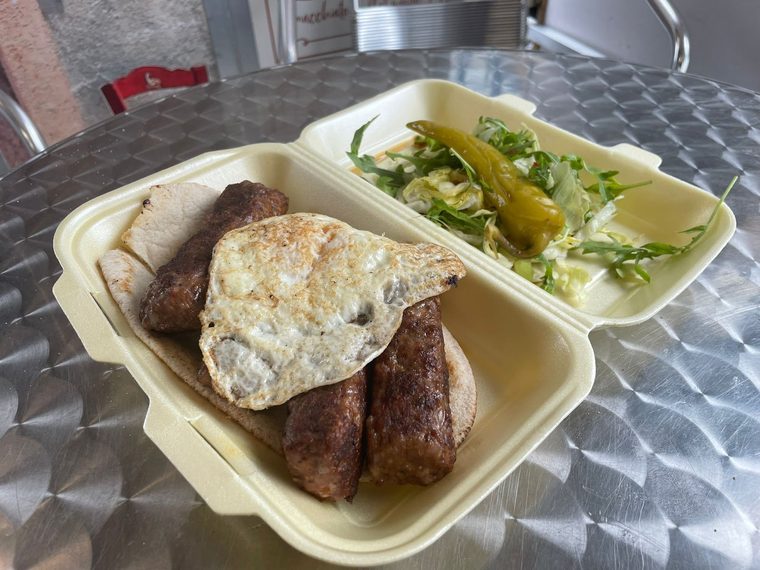 A takeaway box of grilled sausages, fried egg and sliced meat with salad